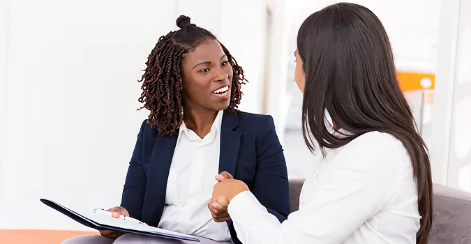 Two businesswomen in discussion, one holding a file, in a bright office setting. Business meeting, collaboration.