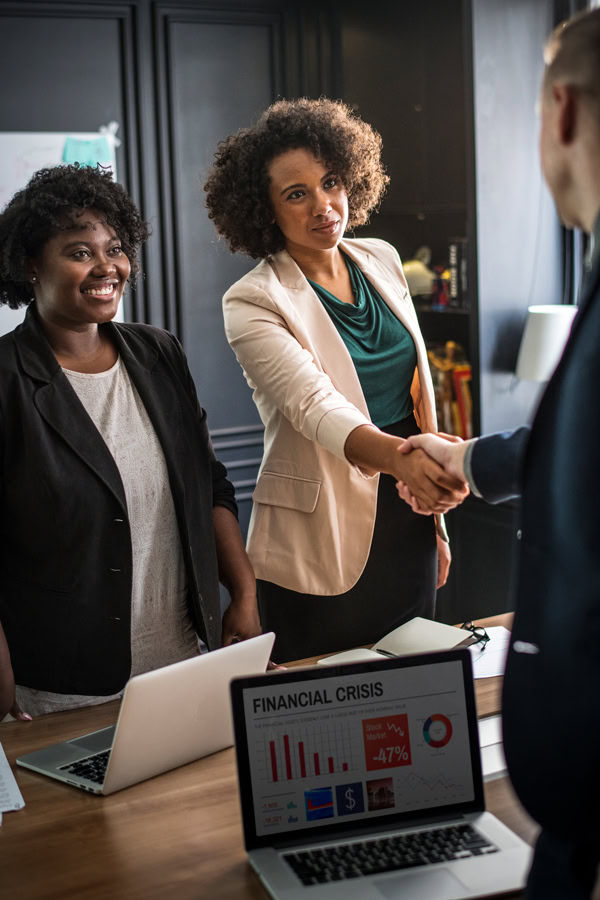 Business meeting: Two African-American businesswomen shaking hands with a colleague. Laptop shows 'Financial Crisis' data.