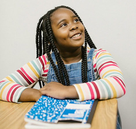 Smiling Black student with braids at school desk. Girl in denim overalls looking up. Education and children concept.